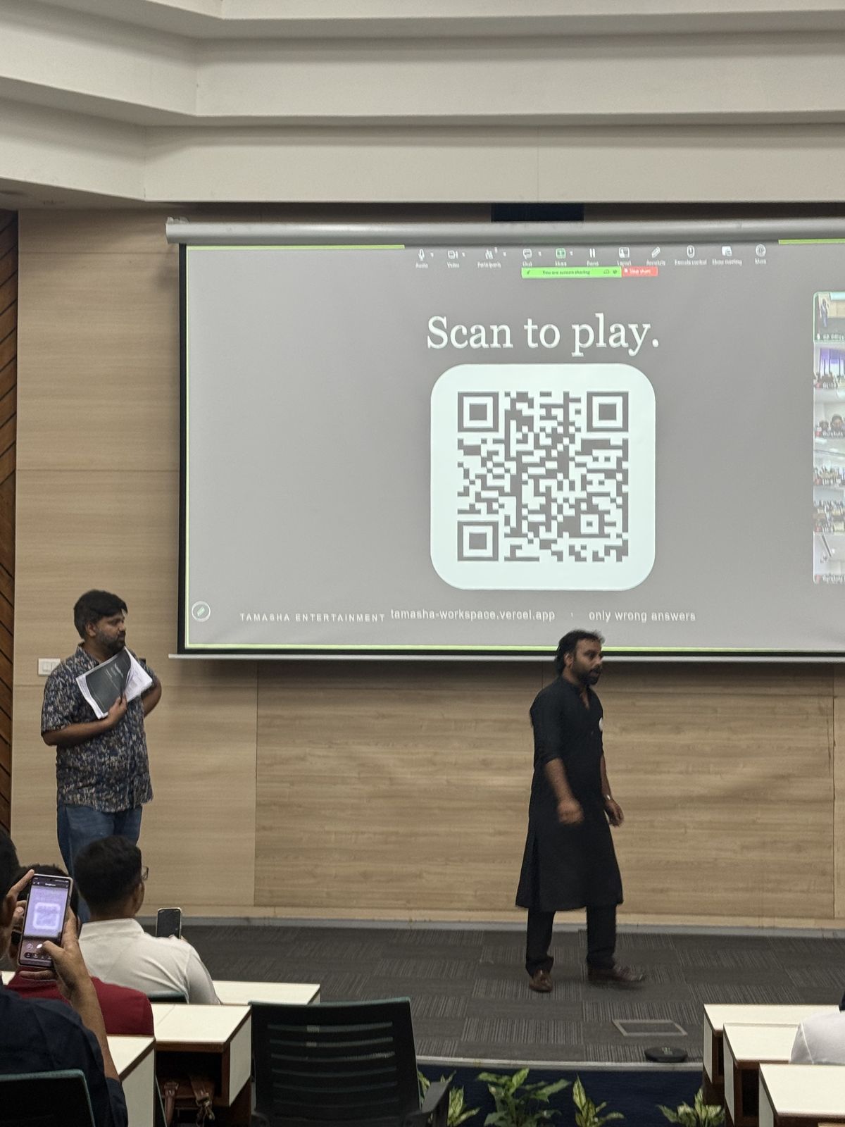A QR code on the big screen in a lecture hall, with two facilitators on stage